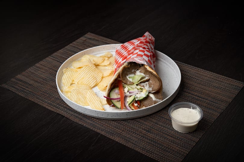Gyro wrap with meat, vegetables, and sauce, served with potato chips and a side of dressing.