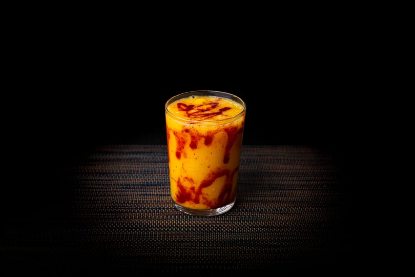 A vibrant yellow mango lassi with red chili sauce swirls in a clear glass.