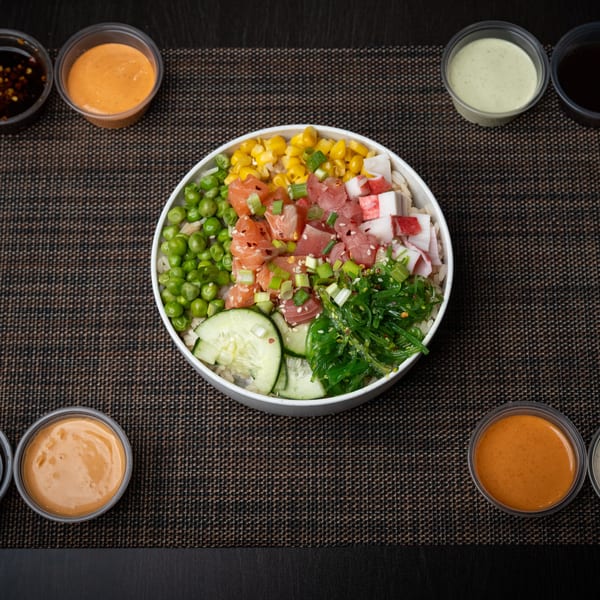 Colorful poke bowl with salmon, tuna, crab, corn, peas, seaweed, and cucumber, surrounded by sauces.