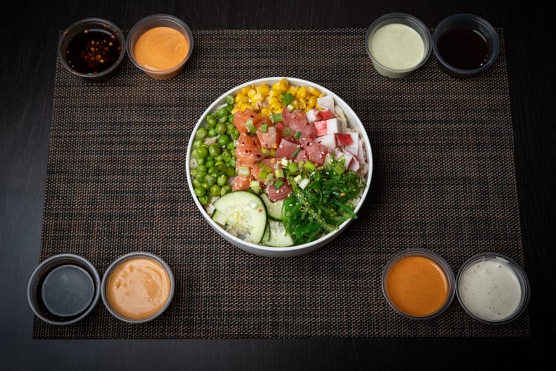 Colorful poke bowl with salmon, tuna, crab, corn, peas, seaweed, and cucumber, surrounded by sauces.