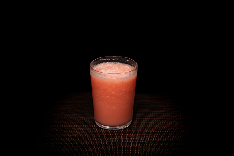 A glass of frothy pink juice sits on a dark, textured surface against a black background.